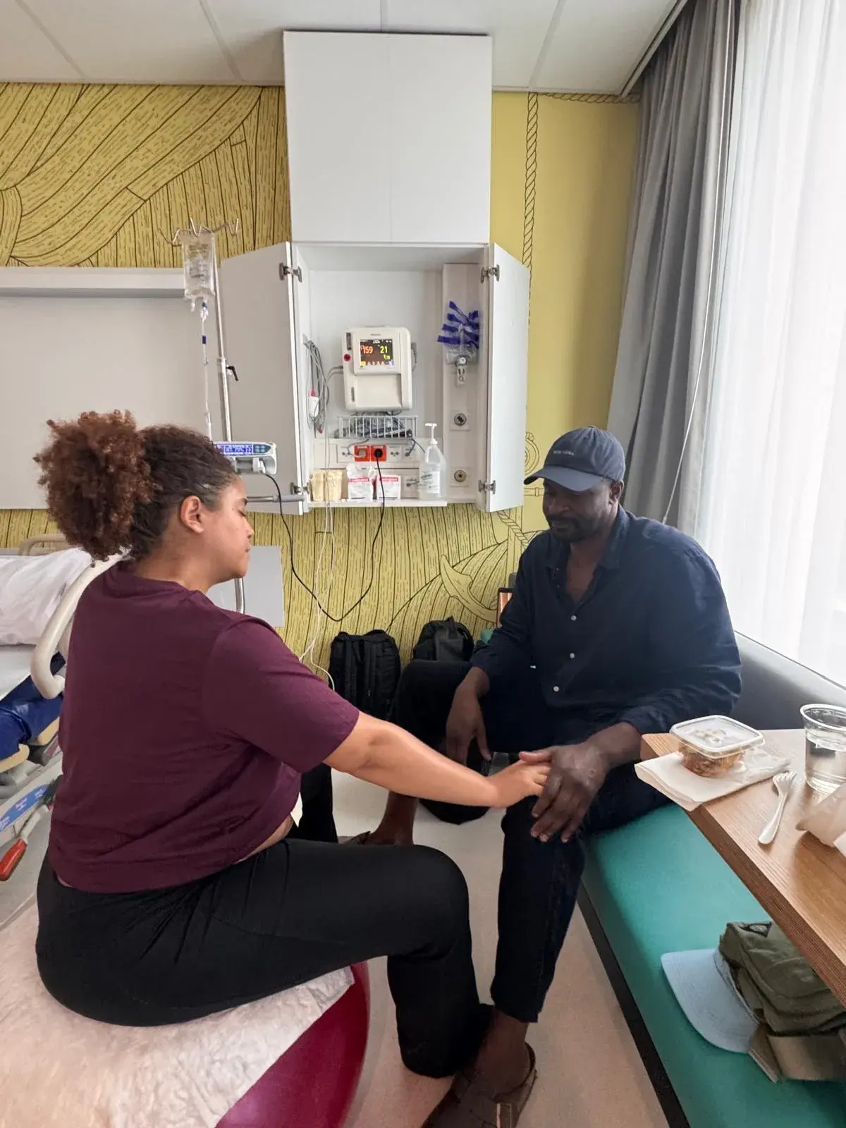Kyle sitting on a birth ball, holding her partner's hands in her hospital room